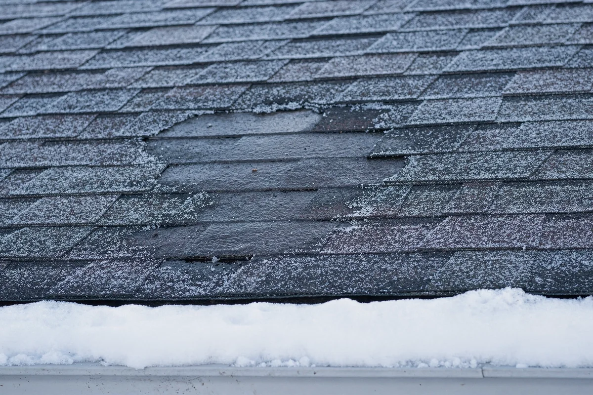 A frosted shingle slope with one bare melted patch, the overhang below still heaped with snow.